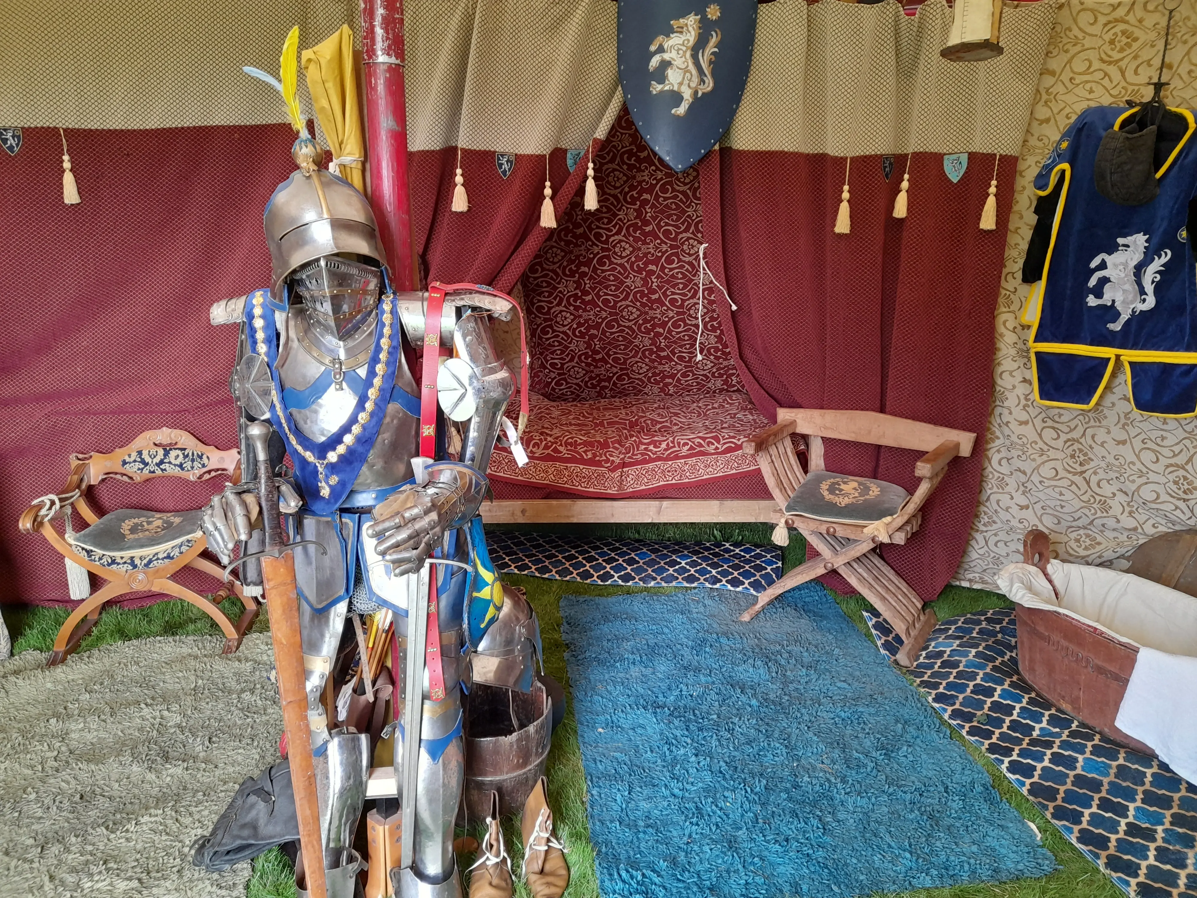 Inside a medieval tent with a suit of armour in the centre, a four-poster bed behind it, wooden chairs on either side, and a wooden bath on the right