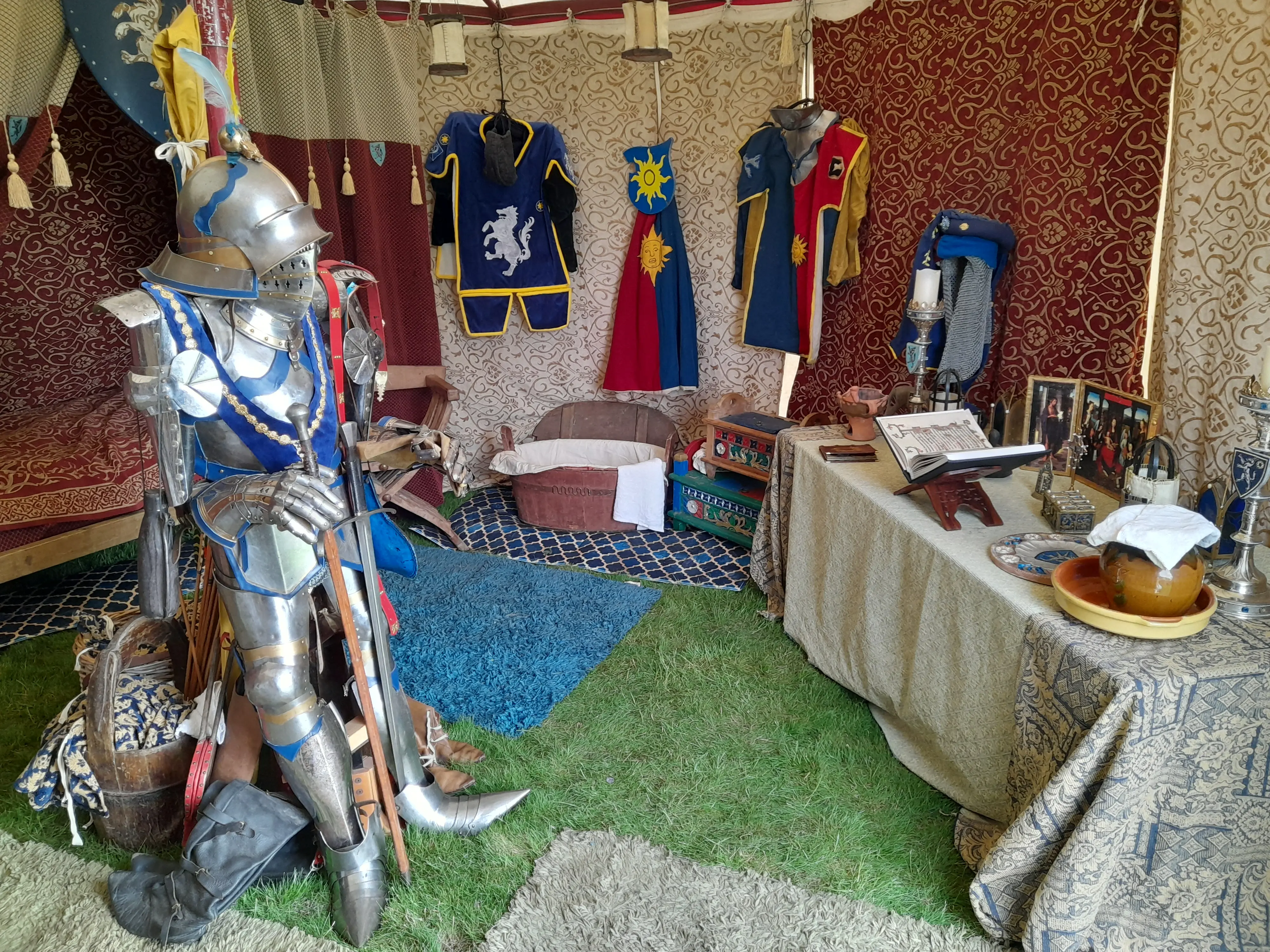 Inside a medieval tent with a suit of armour in the foreground, a four-poster bed to the left, and a religious table right, wooden bath, and clothing at the rear
