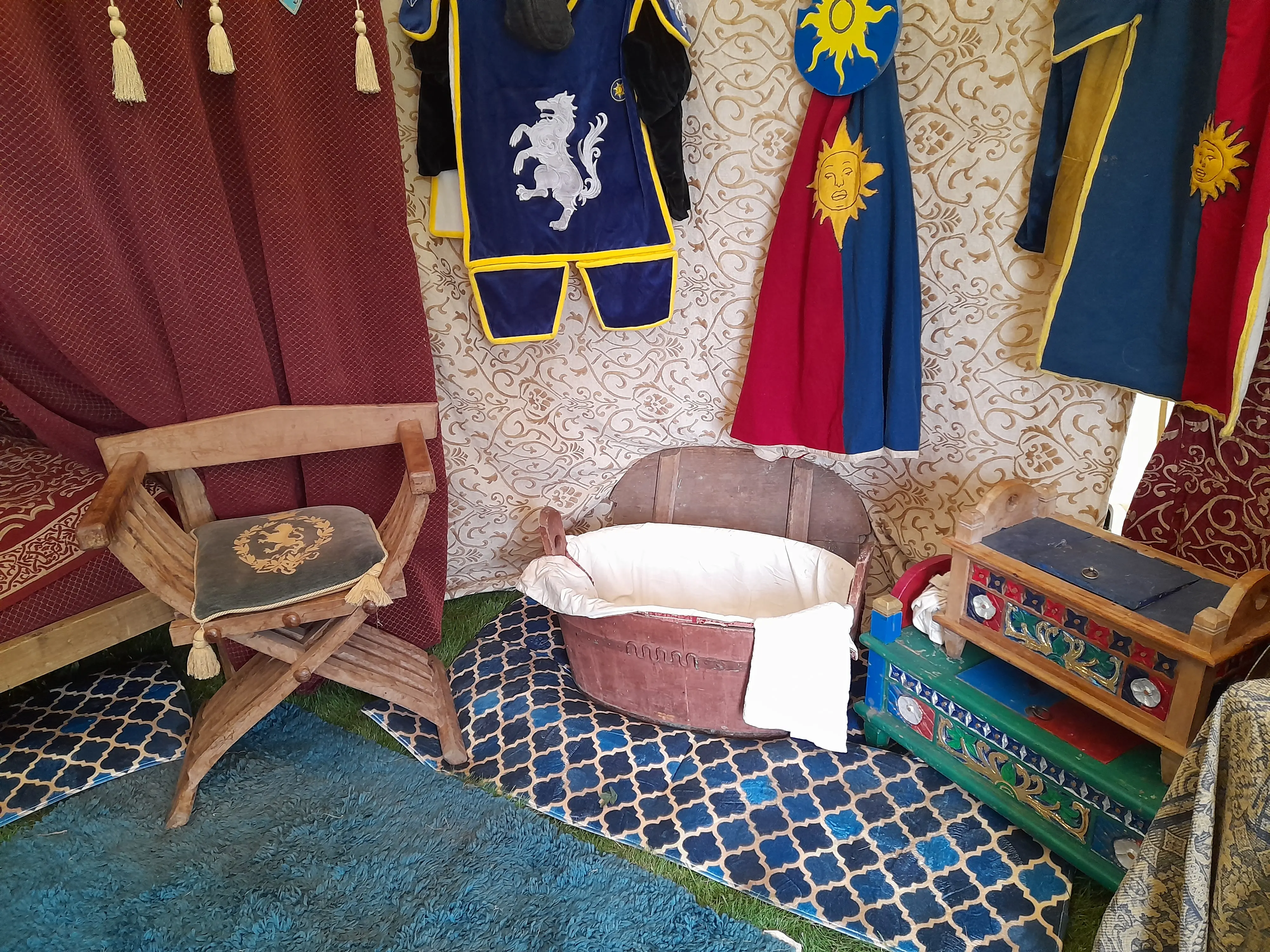 Inside a medieval tent with a wooden chest on the right, and a wooden chair, and a wooden bath in the centre with hanging clothing above