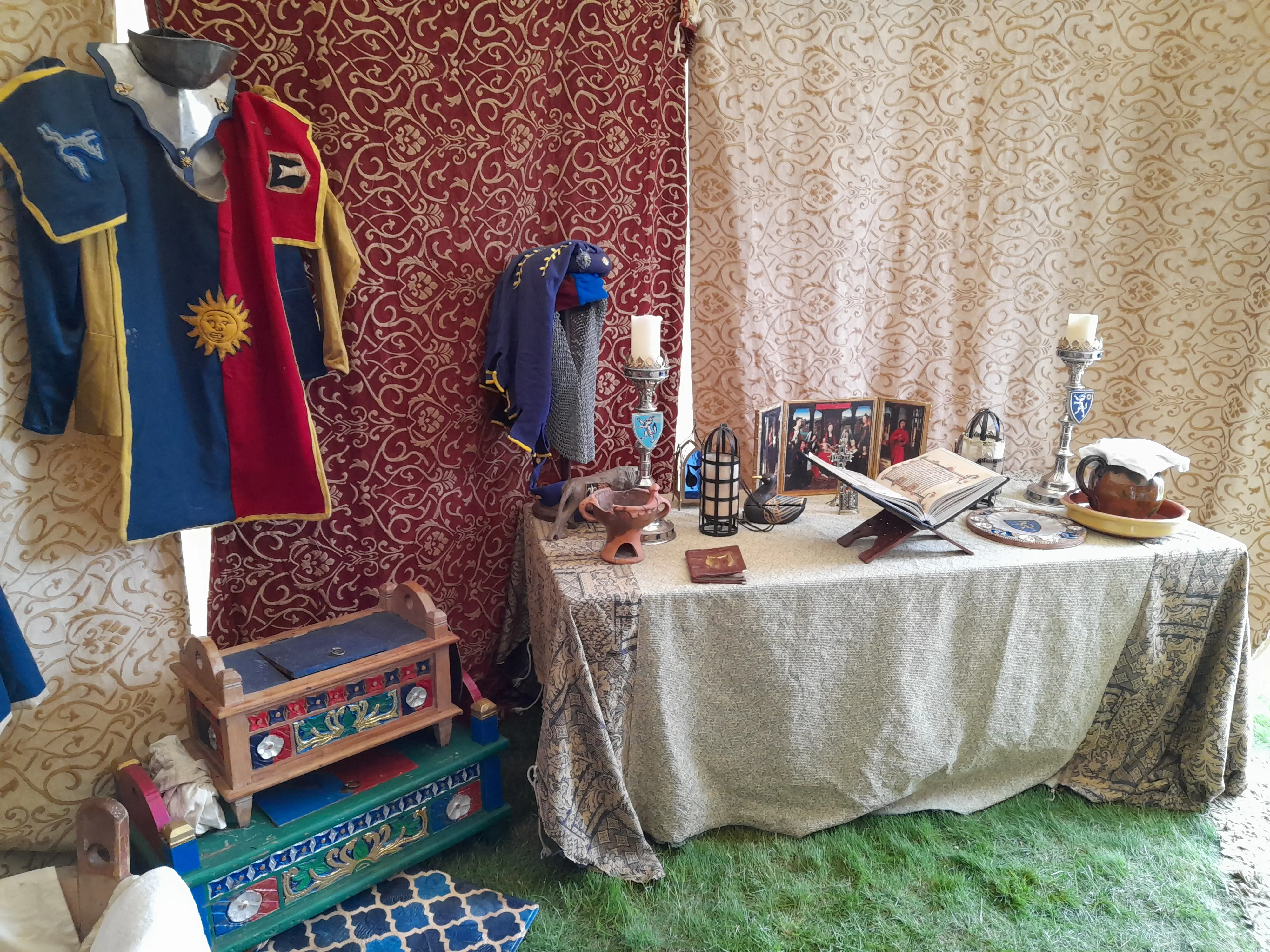 On the left, a hanging medieval surcoat with painted wooden chests below, and a religious table on the centre-right